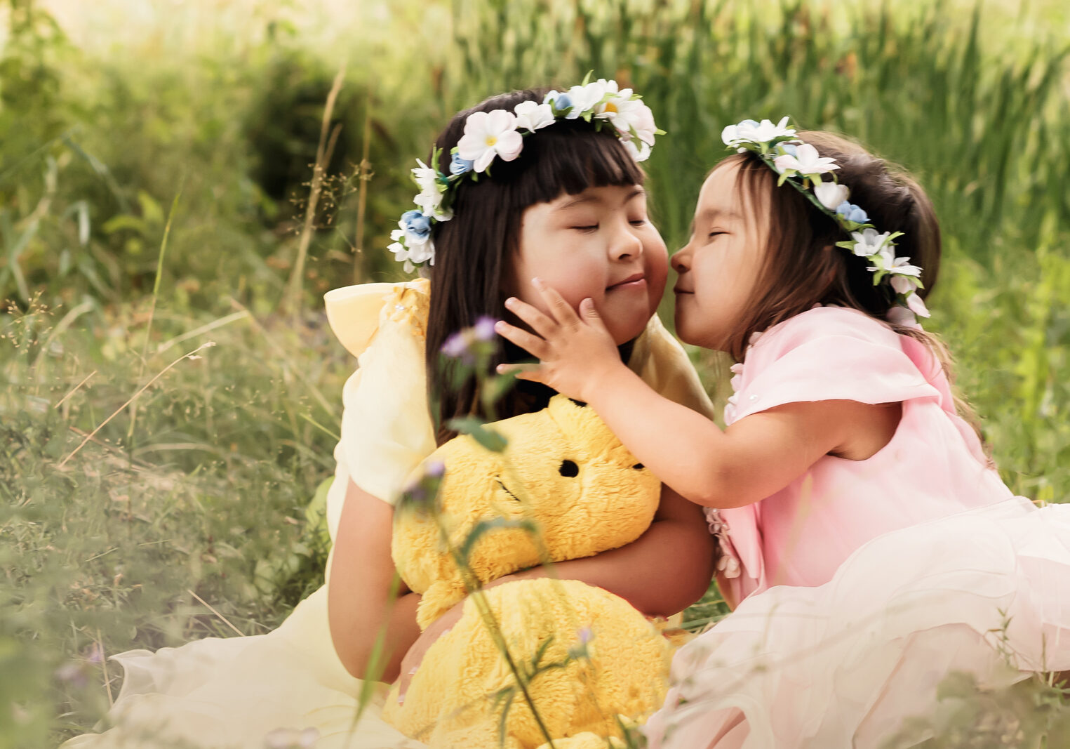 Portrait of two loving sisters wearing princess dresses sitting on a field holding a toy bear.